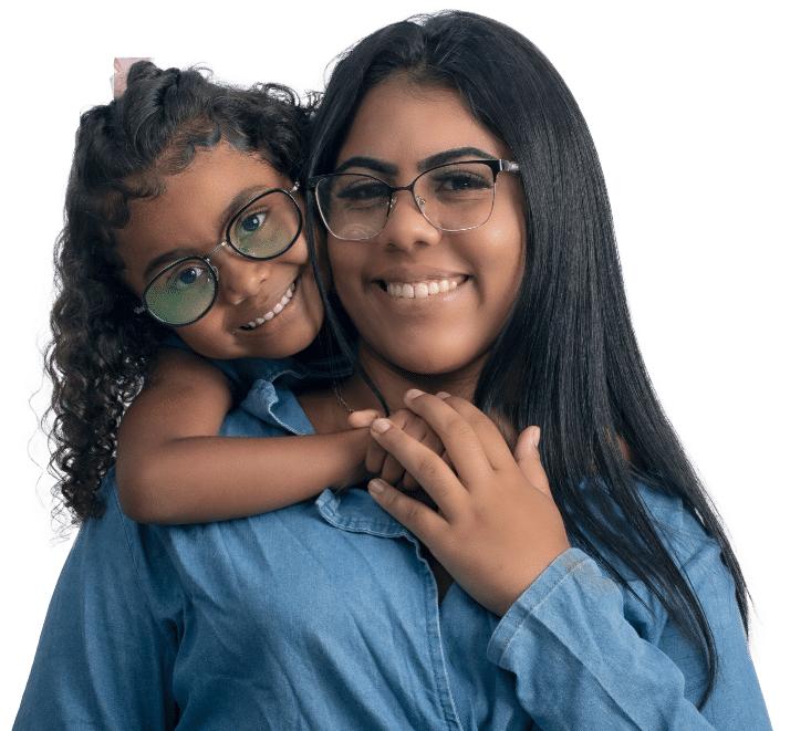 A smiling woman with long dark hair and glasses is hugged from behind by a young girl with curly hair and glasses. They are both wearing blue denim shirts and looking directly at the camera.
