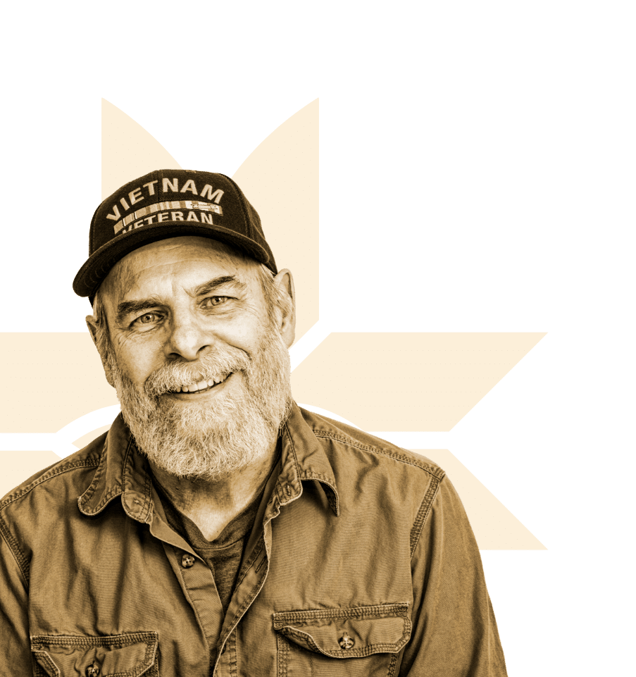 An older man with a gray beard, wearing a Vietnam Veteran cap and a button-up shirt, smiles at the camera. Behind him is a large, yellow star-like graphic on a black background.