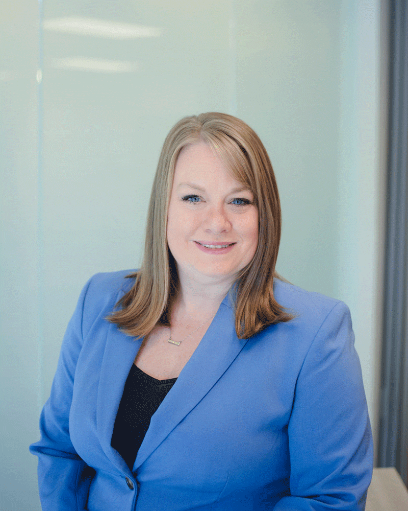 A woman with straight, shoulder-length blonde hair, wearing a blue blazer over a black top, smiles while seated in a bright, modern office setting with glass walls.