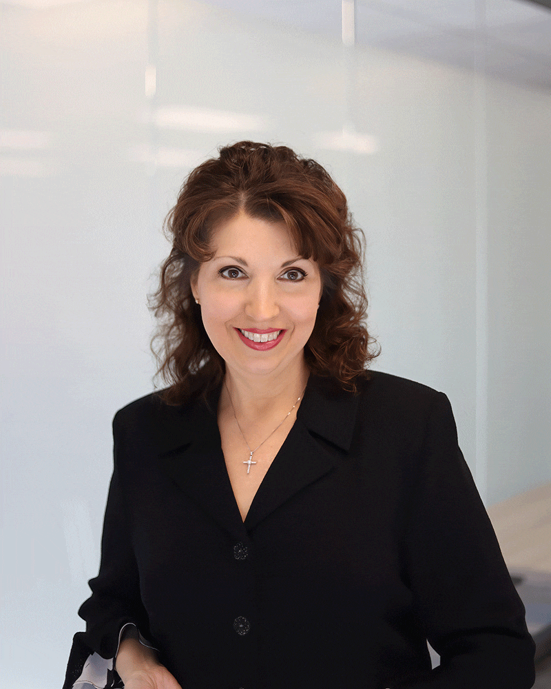 A woman with wavy brown hair, wearing a black blazer and a cross necklace, smiles at the camera in a brightly lit office setting with a glass wall in the background.