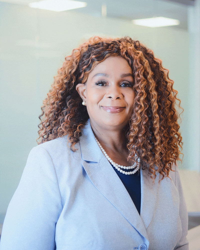 A woman with long, curly hair wearing a light blue blazer, black top, and pearl necklace smiles while standing in a brightly lit office setting.