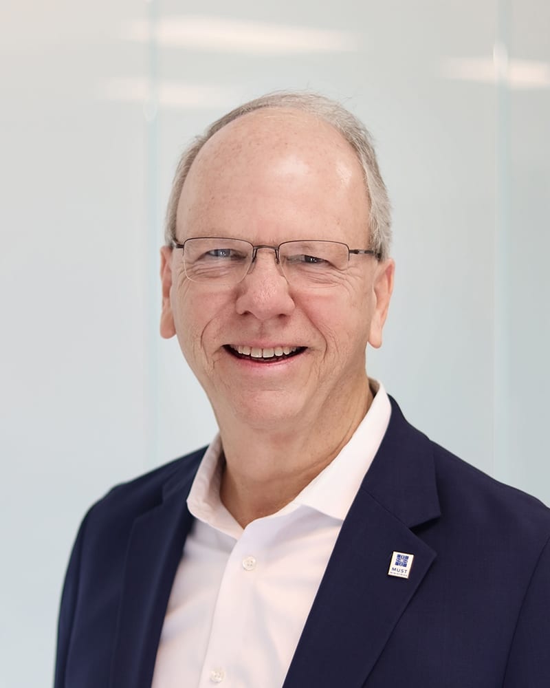 Smiling older man with glasses and short gray hair wearing a navy blazer, white dress shirt, and a pin on his lapel, standing in front of a light, blurred background.
