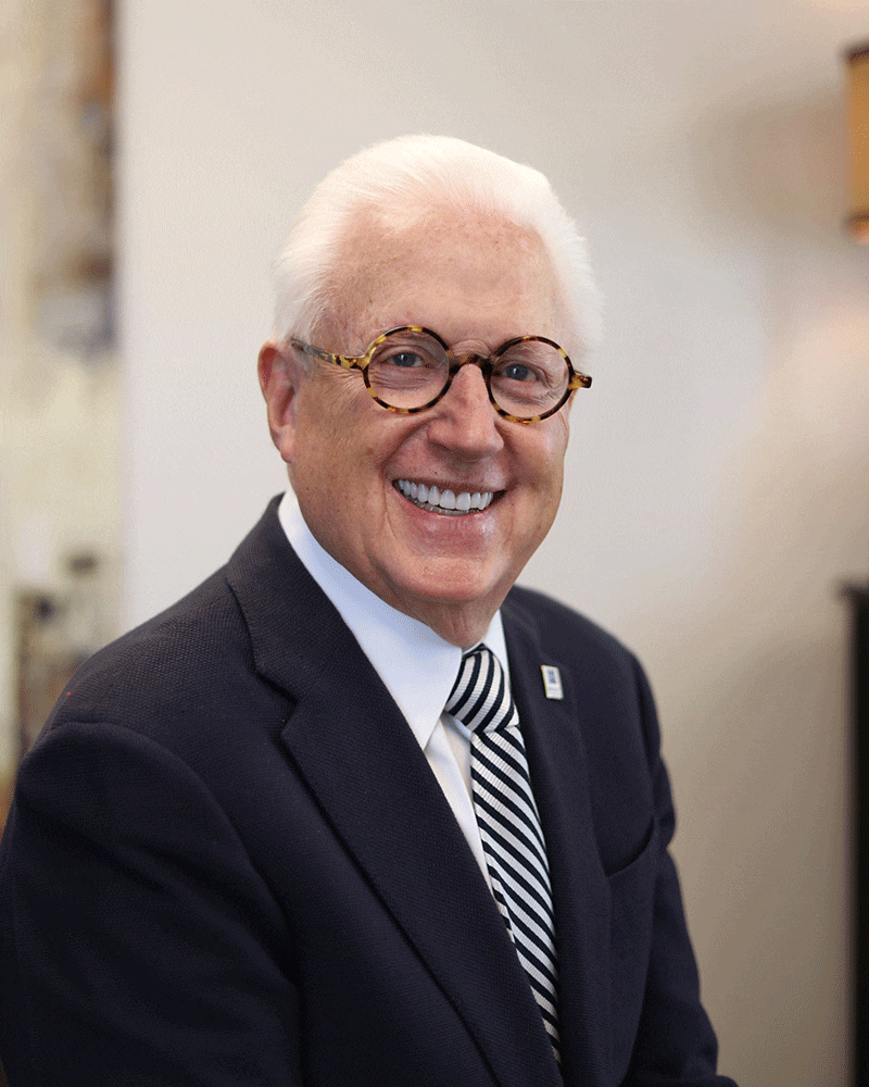 An older man with white hair and round glasses smiles while wearing a dark suit, white shirt, and striped tie. He is posed in an indoor setting with neutral-colored walls in the background.