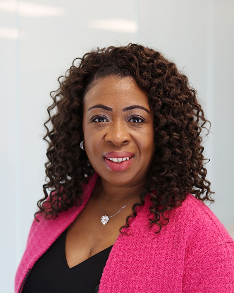 A woman with curly brown hair, wearing a pink textured blazer over a black top and a flower-shaped necklace, smiles at the camera in a brightly lit indoor setting.