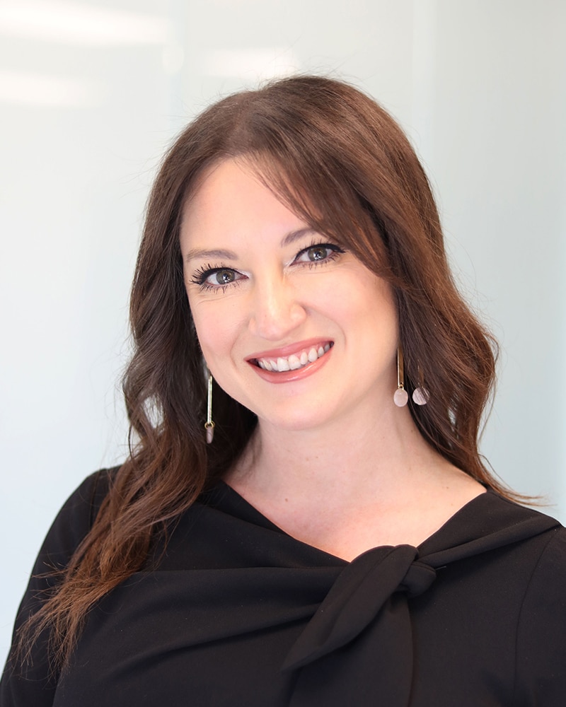 A woman with wavy brown hair wearing a black top and long gold earrings smiles at the camera in a brightly lit, modern setting.