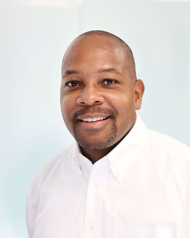 A smiling man wearing a white button-up shirt stands in front of a light blue background. He has short hair, a trimmed goatee, and is looking directly at the camera.