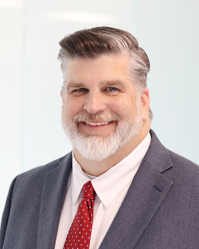 A smiling middle-aged man with gray hair and a beard, wearing a blue suit, white shirt, and red polka dot tie, posed against a light blurred background.