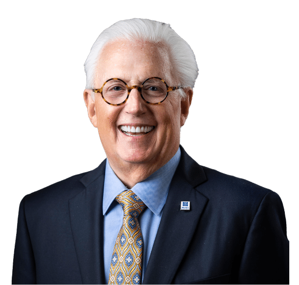 Smiling older man with white hair and glasses wearing a dark suit, light blue shirt, and patterned yellow tie, posing against a black background.