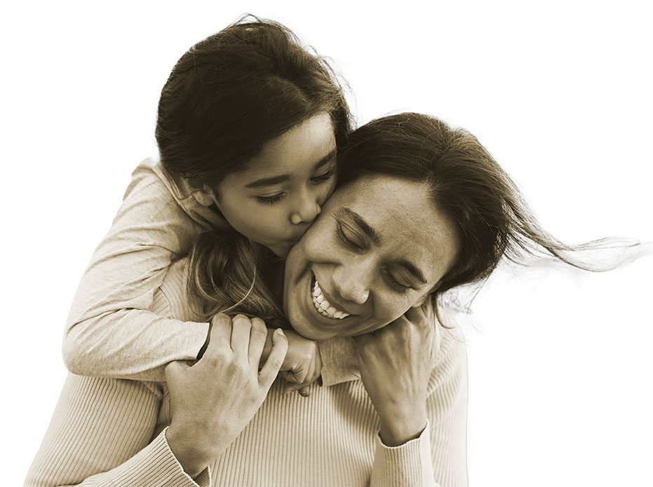 A young girl embraces and kisses a smiling woman on the cheek from behind, both wearing light sweaters, against a plain white background.