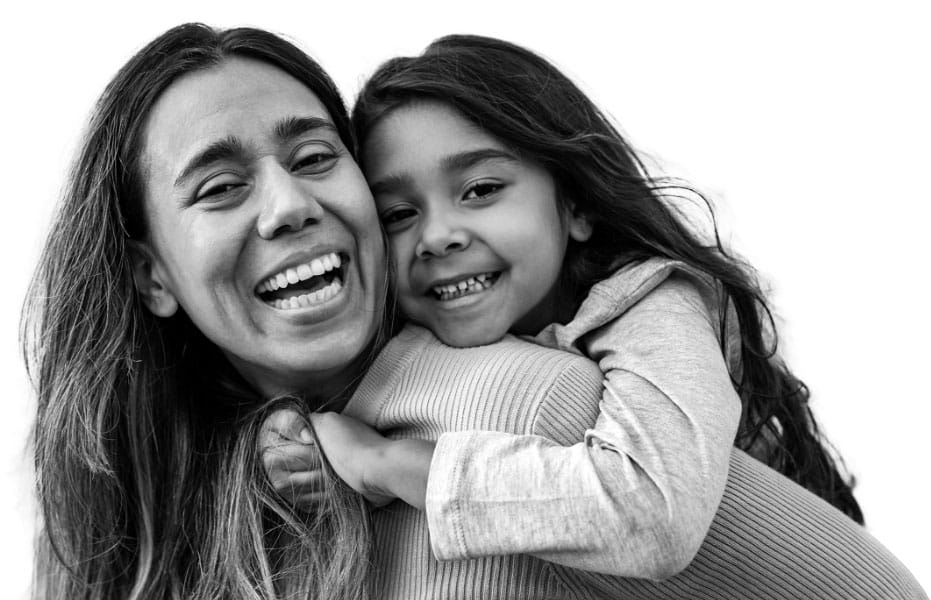 A woman smiles joyfully while a young girl hugs her from behind, both looking at the camera. The image is black and white with a bright, plain background.