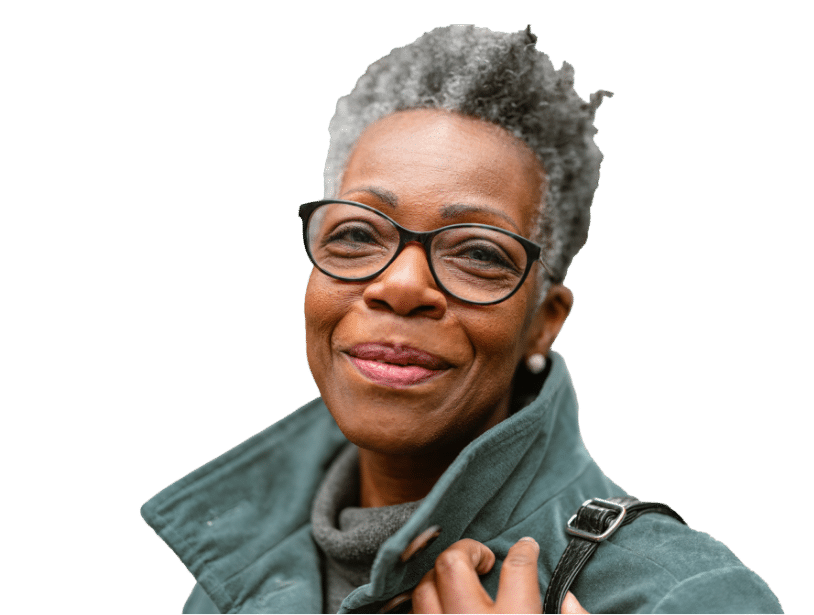 A smiling older woman with short gray hair and glasses wears a green coat and holds the strap of her bag. She looks confidently at the camera against a plain background.