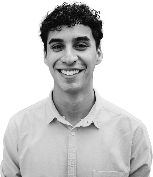 A young man with short curly hair smiles at the camera. He is wearing a light-colored button-up shirt. The background is plain and white. The image is in black and white.