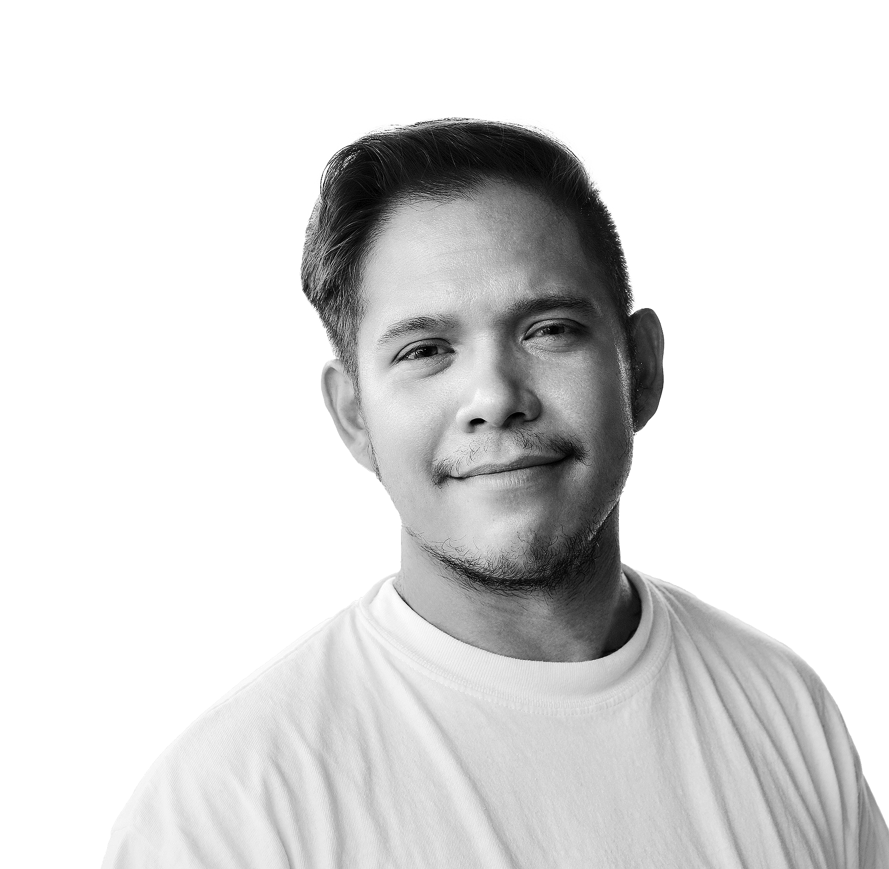 A man with short dark hair and a trimmed beard, wearing a white t-shirt, smiles gently at the camera. The background is blurred with natural light and trees visible. The image is in black and white.