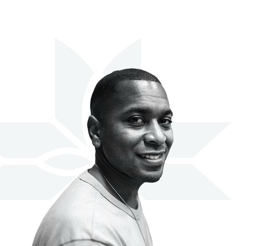 A man with short hair smiles while looking at the camera. He is wearing a light-colored shirt and a necklace. The background is white with a faint, geometric floral design.