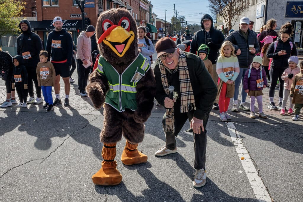 A person in a turkey mascot costume poses with a smiling man in a scarf at a street event, surrounded by people and children wearing race bibs and costumes on a sunny day.