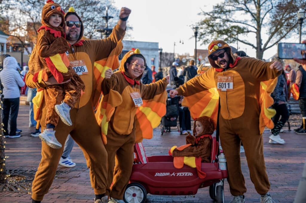 Four people and two children, all dressed in turkey costumes with race bibs, pose cheerfully outdoors. One child is held up, and another sits in a red Radio Flyer wagon. Other people are visible in the background.