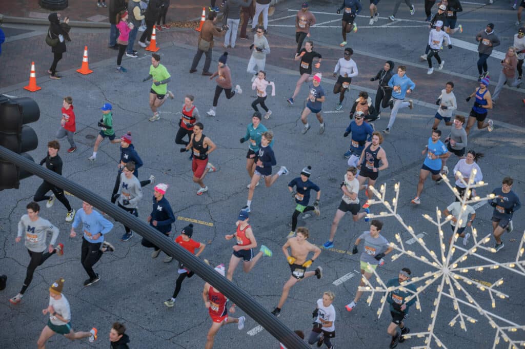 A large group of runners participate in a road race on a city street, seen from above. Some runners wear hats and gloves, indicating cold weather. A decorative, lighted snowflake hangs in the foreground.