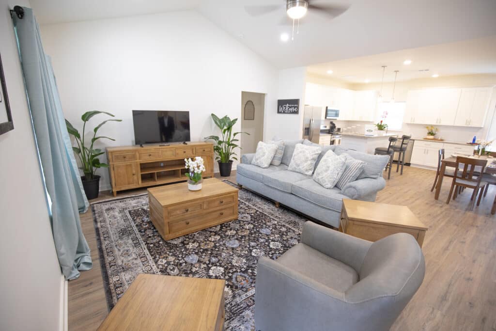 A bright, open living room with a gray sofa, armchair, wooden coffee table, TV stand, large patterned rug, potted plants, and a view into a modern white kitchen and dining area with wood flooring.