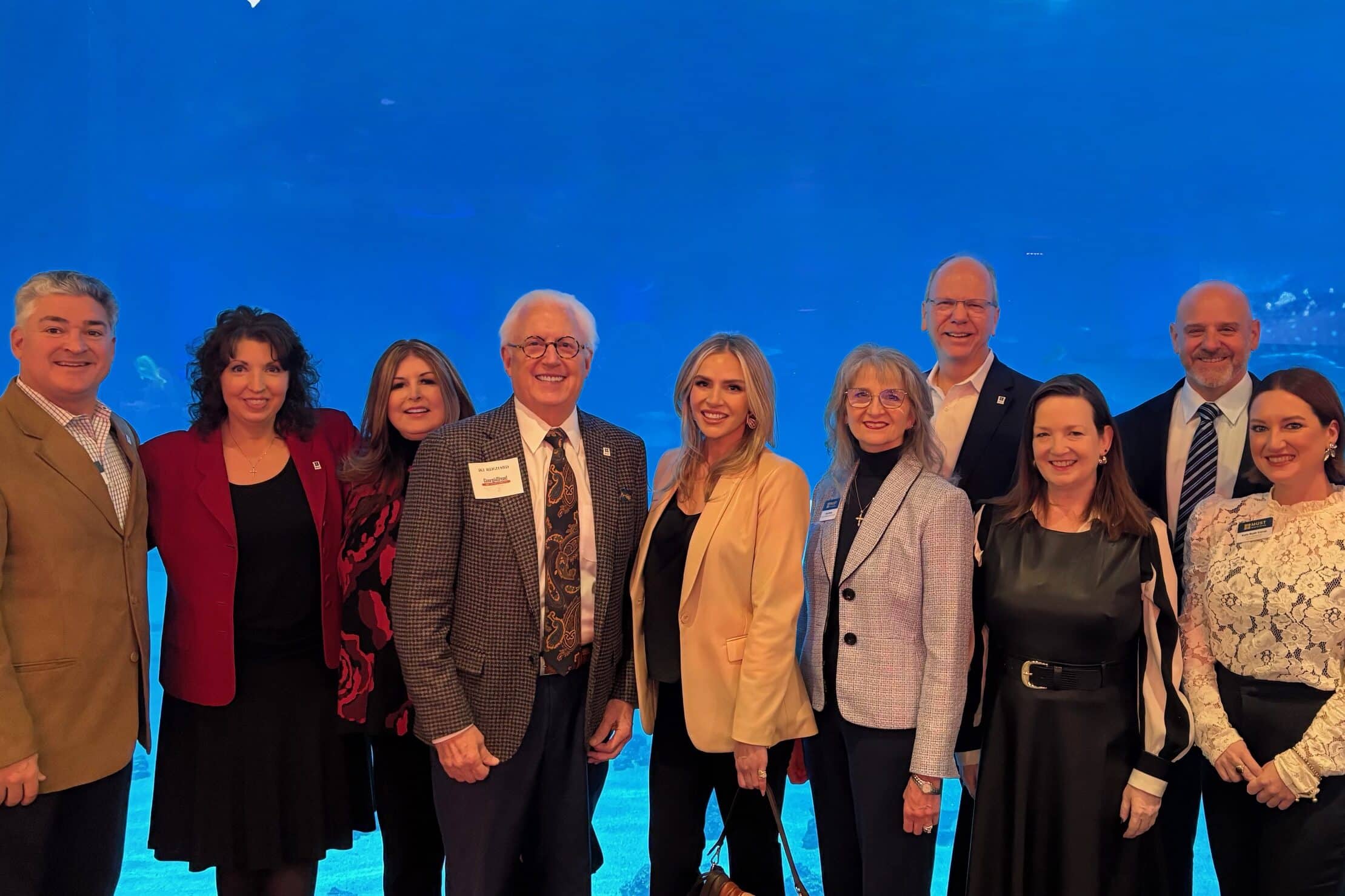 A group of ten adults dressed in business or formal attire stand posing and smiling in front of a large aquarium tank with blue water and visible sea life.