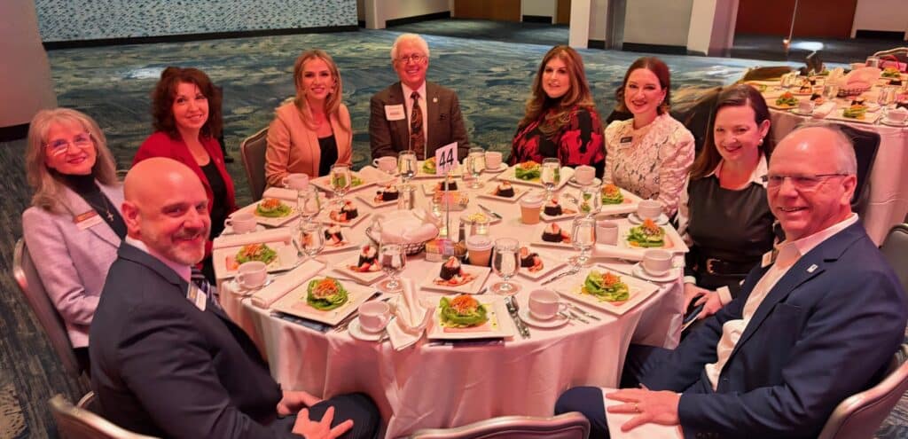 Nine people in business attire are seated around a round table set with salads, drinks, and place settings at a formal event in a banquet hall. Everyone is smiling and looking at the camera.