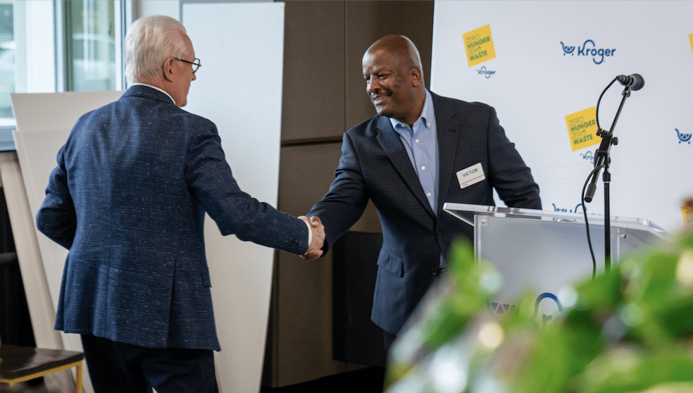 Two men in business attire shake hands at a podium during a formal event, with a microphone and a backdrop displaying the Kroger logo and “Zero Hunger Zero Waste,” highlighting their commitment to addressing food insecurity.