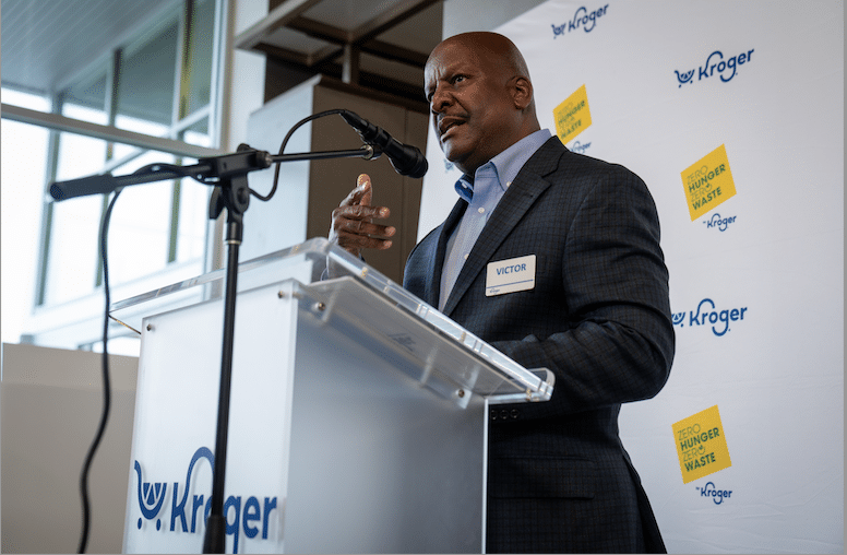 A man in a suit, identified by a badge as Victor, speaks at a podium with a microphone. The Kroger logo and “Zero Hunger | Zero Waste” signs highlight efforts to support food insecure families through neighborhood pantry initiatives.