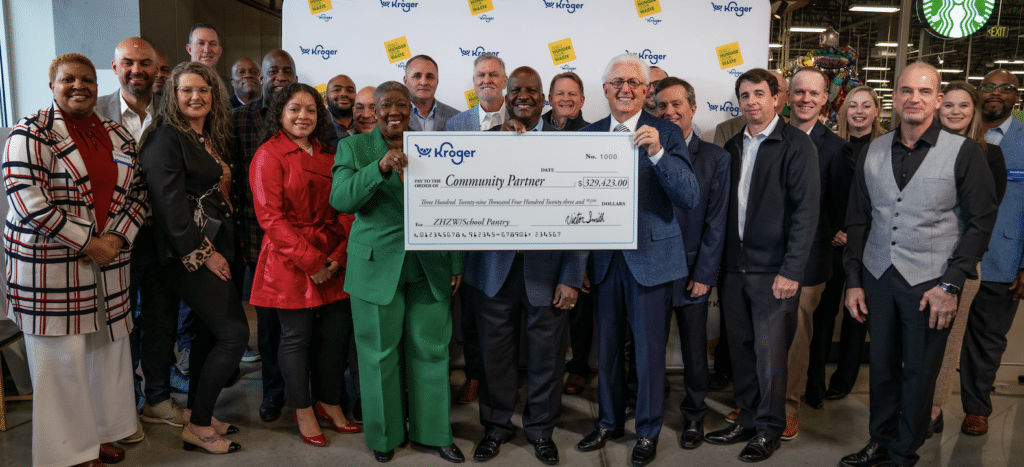 A diverse group smiles and poses indoors, holding an oversized check from Kroger for $287,420.50 to Community Partner in support of Must Ministries’ food donation efforts. Kroger branding and a Starbucks logo are visible in the background.