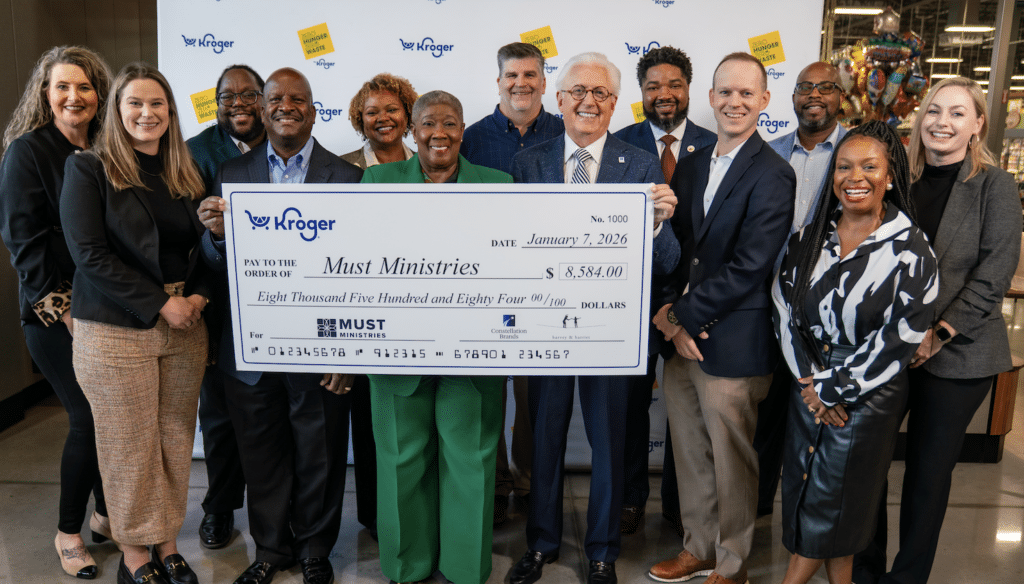 A group of people smile and pose together indoors, holding a large ceremonial check from Kroger for $8,584.00 to Must Ministries, supporting food donation efforts for the food insecure in the community, dated January 7, 2026.