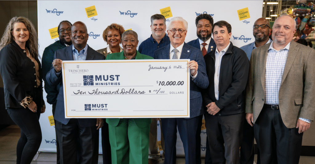 A group of people smile and pose indoors while holding a large $10,000 check from Kroger to MUST Ministries, supporting food insecure families. A Kroger banner is in the background.