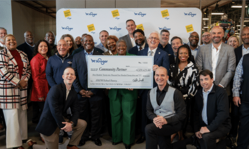 A diverse group of people poses indoors, smiling and holding an oversized Kroger check for a community partner donation. A Starbucks sign is visible in the background.