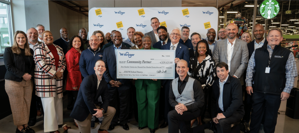 A diverse group of smiling adults poses indoors, some kneeling, holding a large check from Kroger labeled Community Partner to support Must Ministries and those who are food insecure. A Starbucks sign hangs in the background; everyone appears cheerful and celebratory.