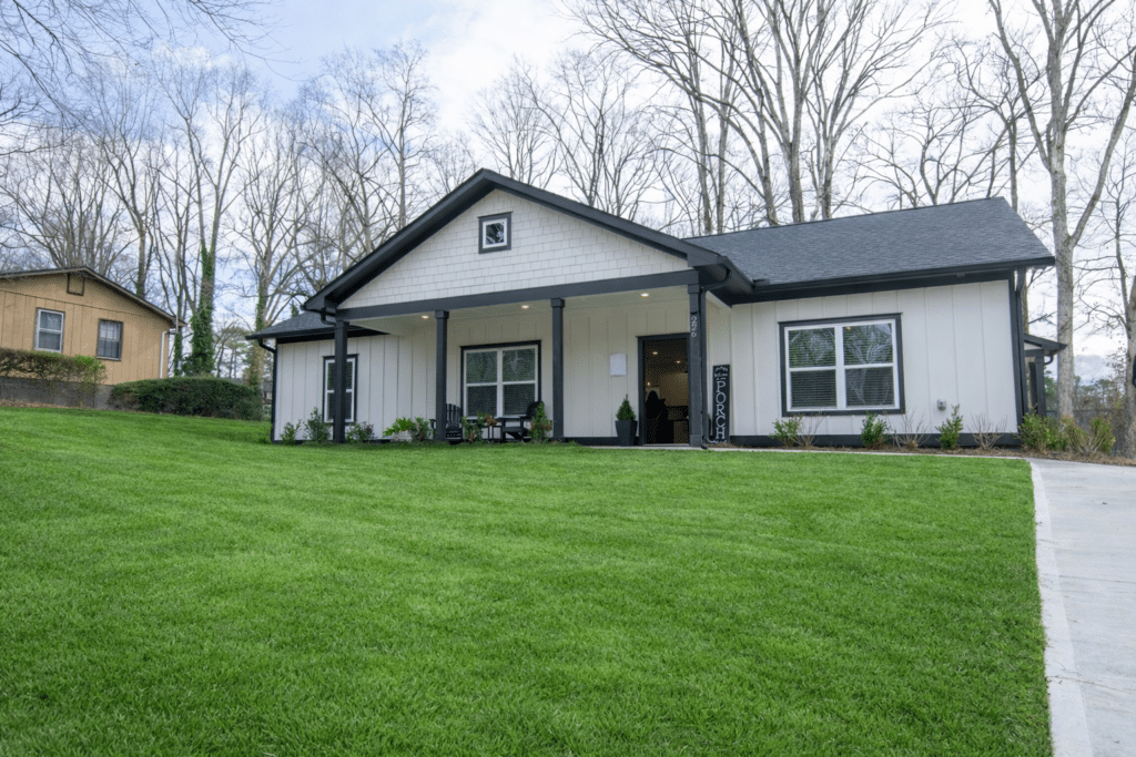 A modern single-story house with white siding and black trim, a covered front porch, and a well-maintained green lawn, set against leafless trees and a cloudy sky.