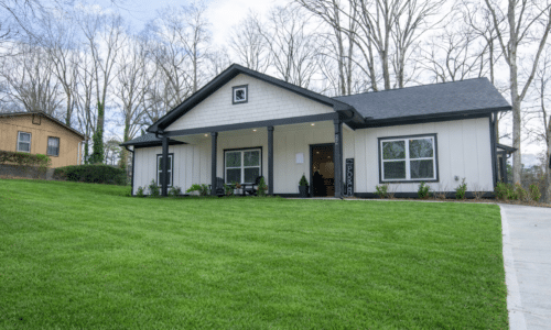 A modern single-story house with white siding and black trim, a covered front porch, and a well-maintained green lawn, set against leafless trees and a cloudy sky.