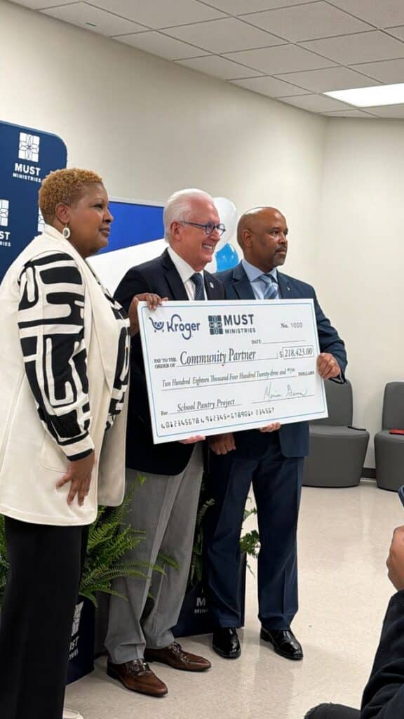 Three people stand together smiling and holding a large ceremonial check from Kroger to MUST Ministries for $218,423, supporting a school pantry project. They are dressed in business attire in a formal indoor setting.