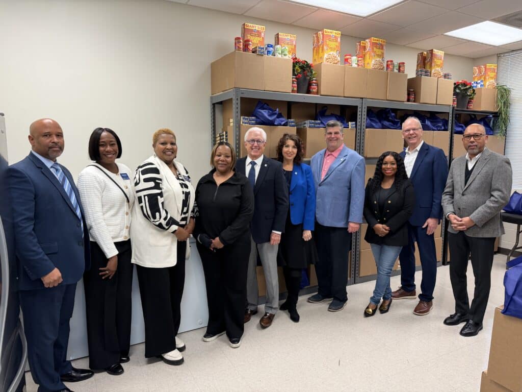 Ten people stand in a row in a room with shelves of food boxes and cereal behind them. They are dressed in business or business-casual attire and are smiling at the camera.