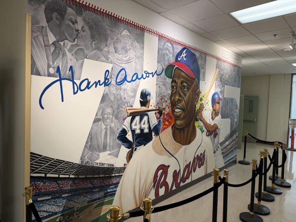 A mural honoring baseball legend Hank Aaron features portraits of him in a Braves uniform, action scenes of him batting, and black-and-white images of cheering fans. A blue signature reads Hank Aaron across the artwork.