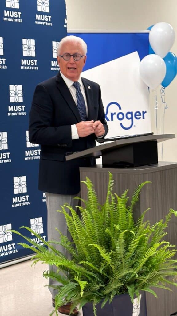 A man in a suit and tie speaks at a podium with a plant in front. Behind him are banners with the logos of MUST Ministries and Kroger, and blue and white balloons are visible to the side.