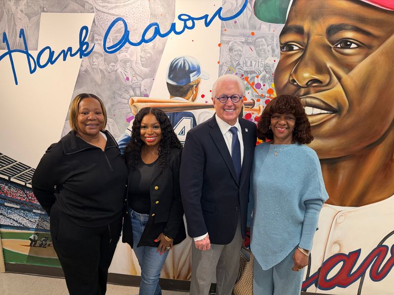 Four people stand smiling in front of a colorful mural featuring Hank Aarons portrait, his name in large blue script, and baseball imagery in the background.