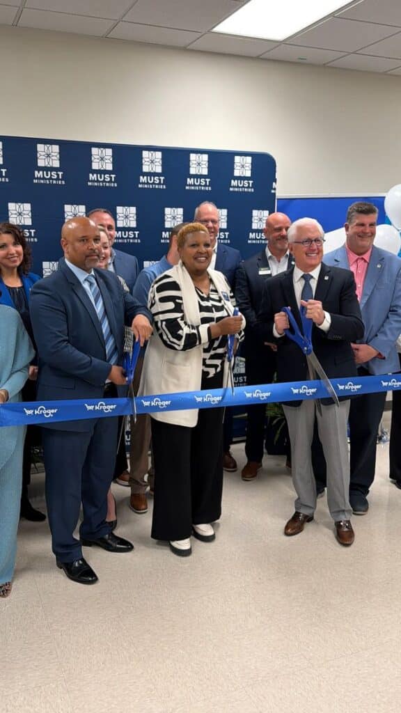 A group of people stand in front of a MUST Ministries backdrop, smiling and holding large scissors to cut a blue ribbon at an indoor ribbon-cutting ceremony. Some wear suits; balloons are visible on the right.