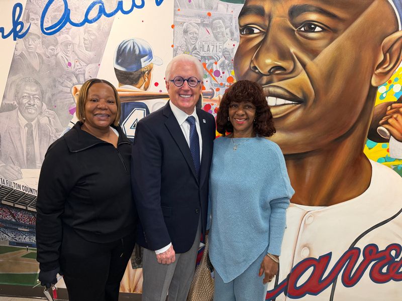 Three people stand smiling in front of a colorful mural featuring a large portrait of a baseball player in a Braves jersey, with other sports-related images in the background.